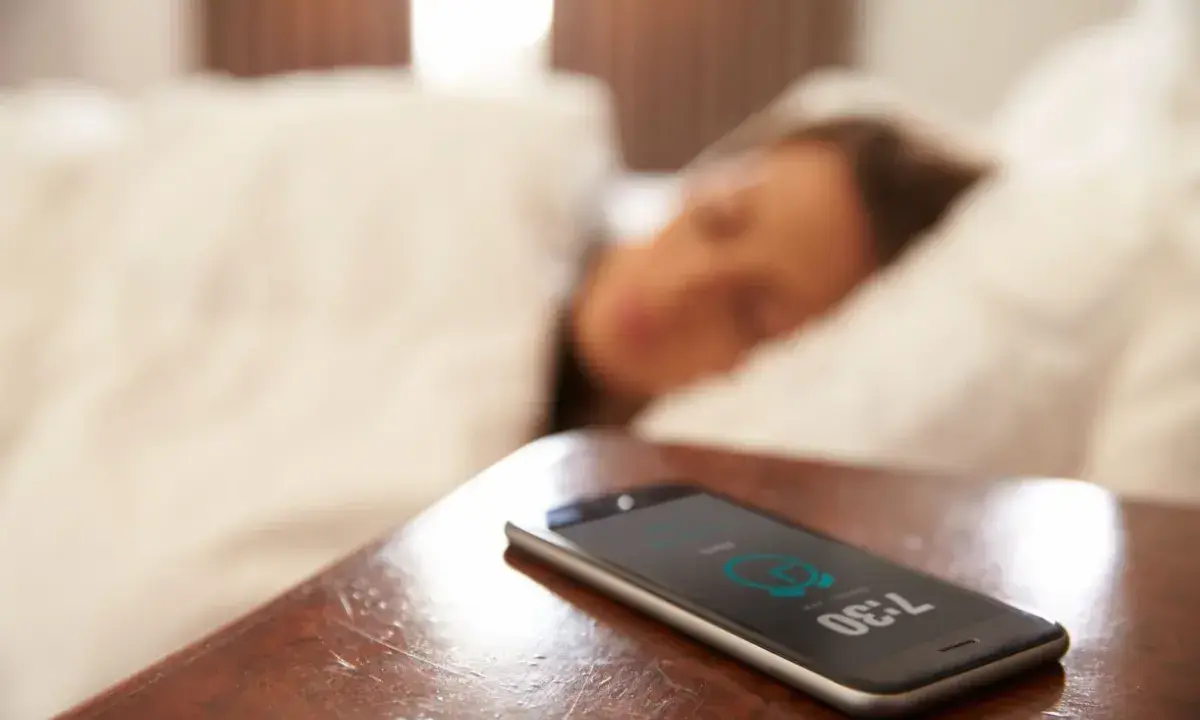 Telefon na stoliku nocnym pokazuje 7:30, budząc śpiącą osobę. To typowy widok na poziomie zero budynku, gdzie zaczyna się dzień.
