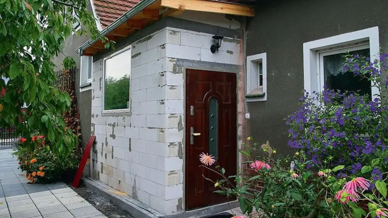 Rozbudowa domu na zgłoszenie: nowe wejście z białych pustaków, brązowe drzwi, okno i kwitnące kwiaty.