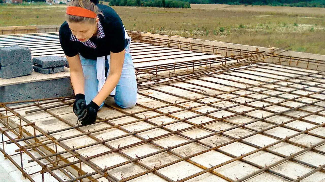 Kobieta w pomarańczowej opasce i jeansach klęczy, wiążąc zbrojenie. To element budowy domu samemu, gdzie każdy detal ma znaczenie.