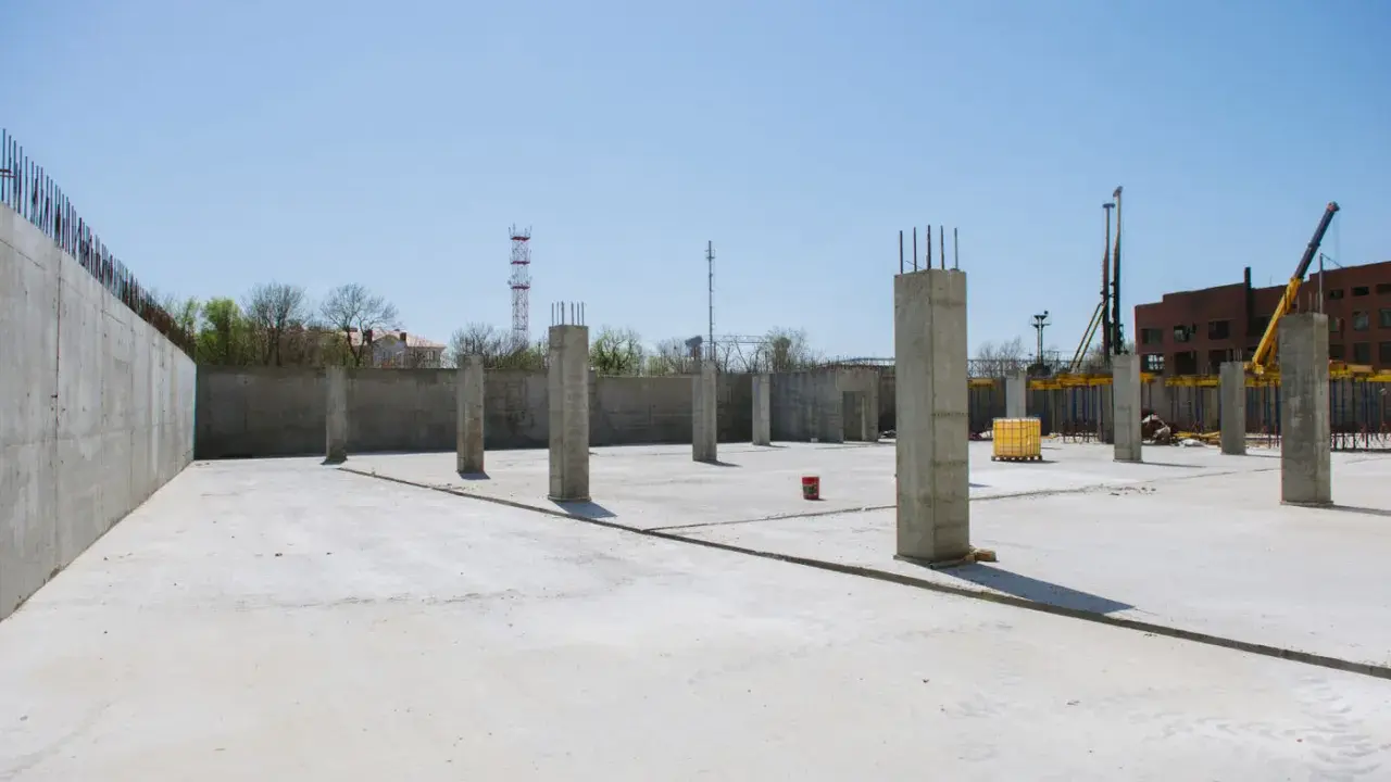 Plac budowy z betonową płytą i słupami betonowymi konstrukcyjnymi. W tle budynek i dźwig.