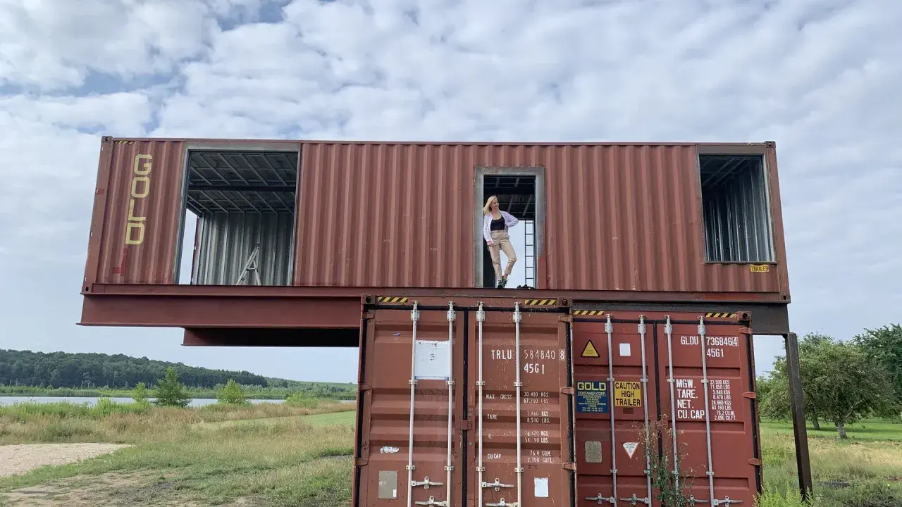 Dom z kontenerów w budowie. Kobieta stoi w otwartych drzwiach górnego kontenera, patrząc na krajobraz.