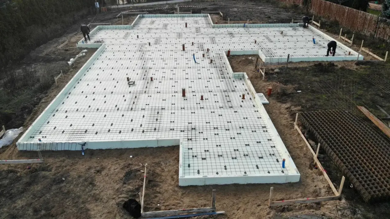 Szalunek płyty fundamentowej gotowy do zalania betonem. Na białej piance izolacyjnej ułożono zbrojenie i rury.