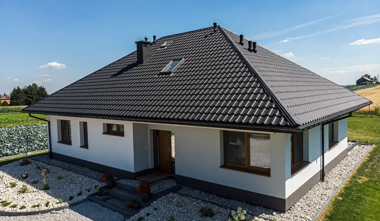 Nowy dom z czarnym dachem, gdzie kalenica jest najwyższym punktem dachu. Wokół zadbany ogród i pola.