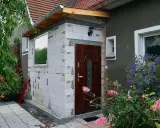 Rozbudowa domu na zgłoszenie: nowe wejście z białych pustaków, brązowe drzwi, okno i kwitnące kwiaty.