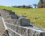 Budowa muru oporowego trwa. Betonowe bloki układane są warstwami, a różowa nić wyznacza linię. Obok stoi drabina.