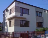 Nowoczesny dom z jasną elewacją ozdobioną poziomymi pasami. Widać kilka okien i fragment ogrodzenia.