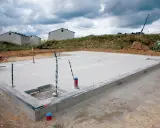 Budowa płyty fundamentowej w trakcie. Betonowa płyta z wystającymi rurami i kablami, w tle domy w budowie.