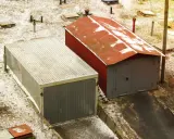 Dwa garaże blaszane, jeden szary z falistym dachem, drugi czerwony z rudym dachem, stoją na zaśnieżonym terenie.