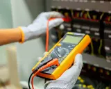 Elektryk w rękawicach wykonuje pomiar instalacji elektrycznej za pomocą żółtego multimetru.