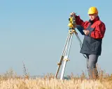 Geodeta w żółtym kasku i czerwonej kurtce pracuje z tachimetrem na polu. Zastanawiasz się, ile bierze geodeta za wytyczenie działki?