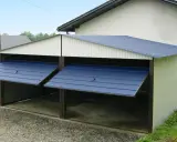 Nowy garaż dwustanowiskowy obok domu, z niebieskimi, uchylnymi bramami. Świetny pomysł na garaż obok domu, zapewniający ochronę pojazdom.