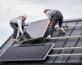 Dwóch monterów instaluje panele fotowoltaiczne na ciemnym, szczelnym dachu. Praca nad przyszłością energetyki.