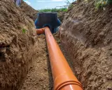 Wykonanie przyłącza wodociągowego: pomarańczowa rura biegnie przez wykop w ziemi do czarnej studzienki.