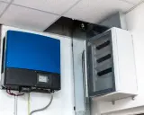 Niebieski falownik, kluczowy element instalacji fotowoltaicznej, zamontowany obok białej skrzynki z bezpiecznikami.