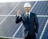 Biznesmen w kasku i garniturze pokazuje znak OK na tle paneli fotowoltaicznych. Odpowiedź na pytanie: czy fotowoltaika się opłaca? Tak!