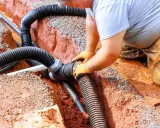 Instalator w pomarańczowych rękawiczkach łączy rury odprowadzające wodę z rynien. Prace zgodne z przepisami.