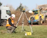 Geodeta w kamizelce odblaskowej wykonuje badanie gruntu pod budowę domu. W tle koparka i ciężarówka.