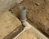 System kanalizacyjny w wykopie. Szara rura do kanalizacji pionowo i poziomo, połączona z innymi elementami.