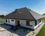 Nowy dom z czarnym dachem, gdzie kalenica jest najwyższym punktem dachu. Wokół zadbany ogród i pola.