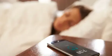 Telefon na stoliku nocnym pokazuje 7:30, budząc śpiącą osobę. To typowy widok na poziomie zero budynku, gdzie zaczyna się dzień.