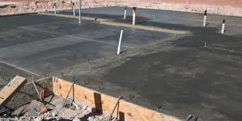 Świeżo wylany betonowy fundament z widocznymi rurami i zbrojeniem. Ziemia wokół czeka na materiał, czym obsypać fundamenty z zewnątrz.