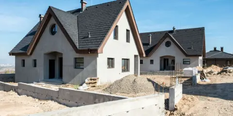 Budowa budynku gospodarczego w toku. Dwa domy z ciemnymi dachami i jasnymi ścianami, w otoczeniu placu budowy z betonowymi fundamentami i gruzem.