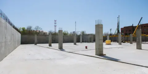 Plac budowy z betonową płytą i słupami betonowymi konstrukcyjnymi. W tle budynek i dźwig.
