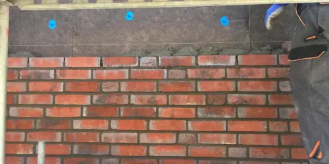 Budowa elewacji z klinkieru. Cegły układane na zaprawie, widoczna ręka w rękawicy i rusztowanie.