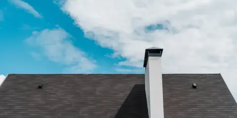 Biały komin systemowy na ciemnym dachu pod błękitnym niebem z chmurami.