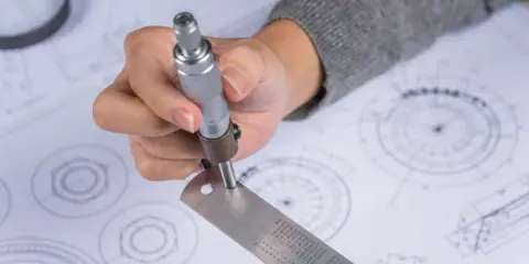 Ręka trzymająca suwmiarkę nad rysunkiem technicznym, pokazując jak czytać rysunek techniczny.