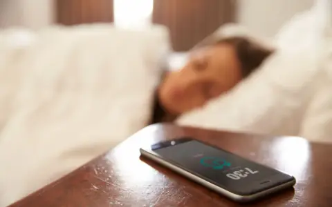 Telefon na stoliku nocnym pokazuje 7:30, budząc śpiącą osobę. To typowy widok na poziomie zero budynku, gdzie zaczyna się dzień.