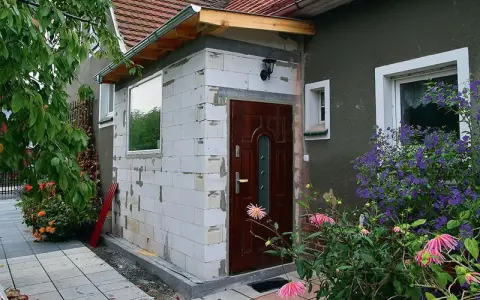 Rozbudowa domu na zgłoszenie: nowe wejście z białych pustaków, brązowe drzwi, okno i kwitnące kwiaty.