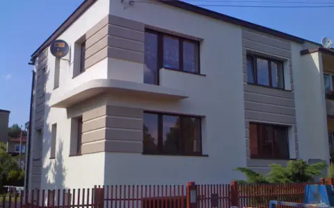 Nowoczesny dom z jasną elewacją ozdobioną poziomymi pasami. Widać kilka okien i fragment ogrodzenia.