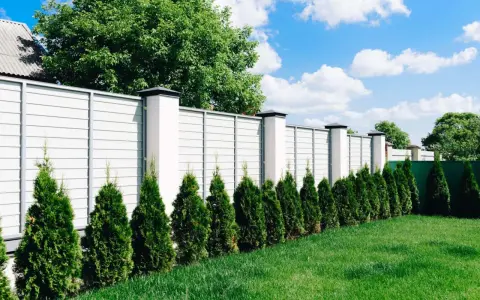 Nowy, biały płot z betonowymi słupkami, otaczający posesję przy drodze gminnej. Rząd zielonych żywotników tworzy naturalną barierę.