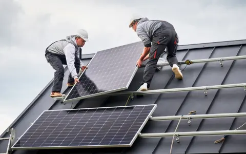 Dwóch monterów instaluje panele fotowoltaiczne na ciemnym, szczelnym dachu. Praca nad przyszłością energetyki.