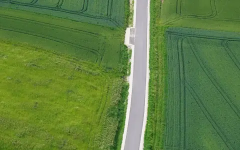 Wąska droga dojazdowa do działki, otoczona polami zielonych zbóż i traw.