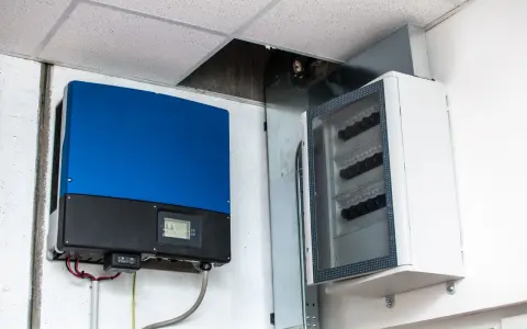 Niebieski falownik, kluczowy element instalacji fotowoltaicznej, zamontowany obok białej skrzynki z bezpiecznikami.