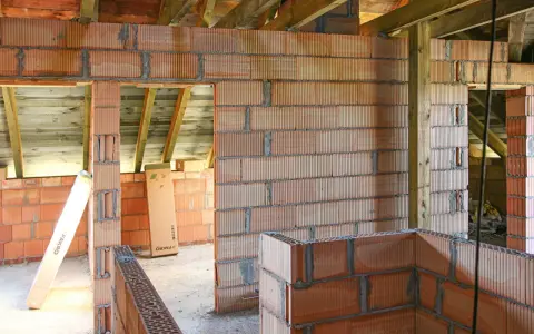 Niedokończone ściany z pustaków ceramicznych, widoczne drewniane belki stropowe. Wady murowania na piankę mogą prowadzić do problemów.