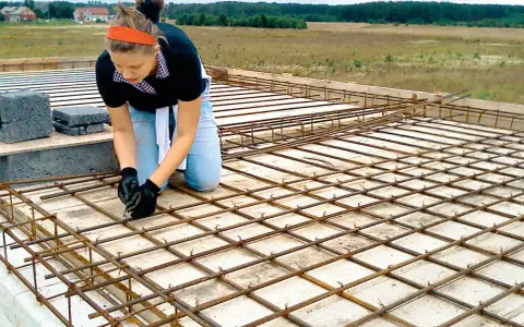 Kobieta w pomarańczowej opasce i jeansach klęczy, wiążąc zbrojenie. To element budowy domu samemu, gdzie każdy detal ma znaczenie.