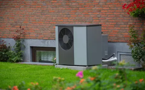 Solidny fundament pod pompę ciepła zapewnia stabilność urządzenia. Urządzenie stoi na trawniku przy ceglanym domu.