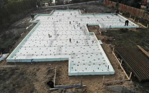 Szalunek płyty fundamentowej gotowy do zalania betonem. Na białej piance izolacyjnej ułożono zbrojenie i rury.
