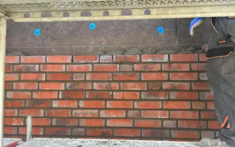 Budowa elewacji z klinkieru. Cegły układane na zaprawie, widoczna ręka w rękawicy i rusztowanie.