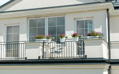 Białe okna pięknie komponują się z jasną, tynkowaną elewacją, tworząc elegancki wygląd. Balkon ozdobiony kwiatami dodaje uroku.