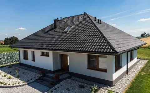 Nowy dom z czarnym dachem, gdzie kalenica jest najwyższym punktem dachu. Wokół zadbany ogród i pola.