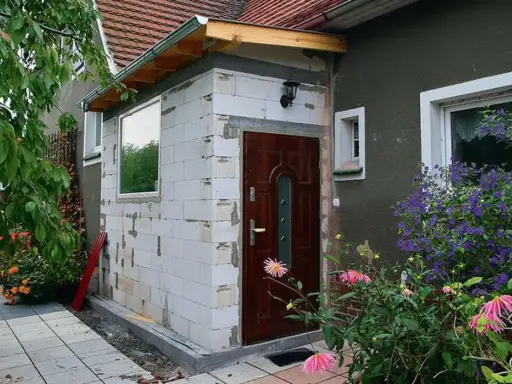 Rozbudowa domu na zgłoszenie: nowe wejście z białych pustaków, brązowe drzwi, okno i kwitnące kwiaty.