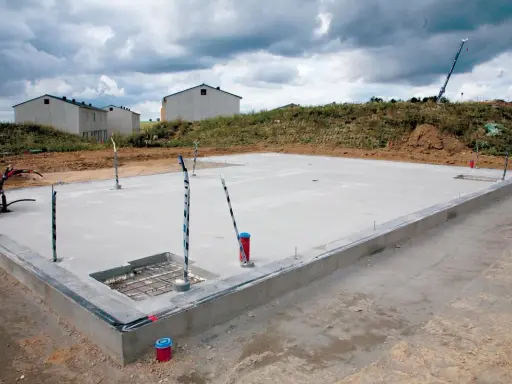 Budowa płyty fundamentowej w trakcie. Betonowa płyta z wystającymi rurami i kablami, w tle domy w budowie.