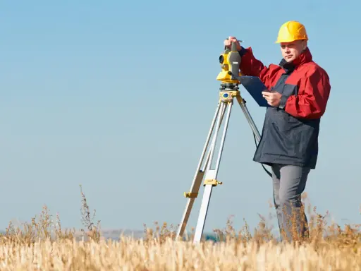 Geodeta w żółtym kasku i czerwonej kurtce pracuje z tachimetrem na polu. Zastanawiasz się, ile bierze geodeta za wytyczenie działki?