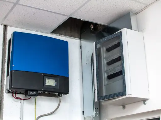 Niebieski falownik, kluczowy element instalacji fotowoltaicznej, zamontowany obok białej skrzynki z bezpiecznikami.
