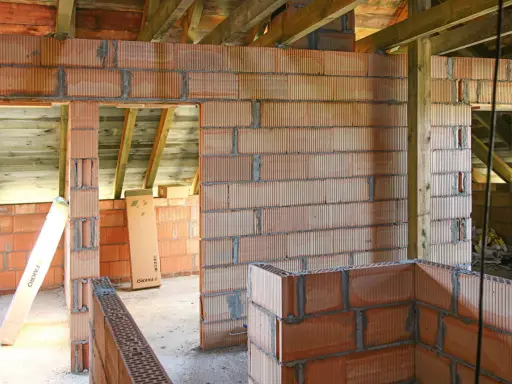 Niedokończone ściany z pustaków ceramicznych, widoczne drewniane belki stropowe. Wady murowania na piankę mogą prowadzić do problemów.