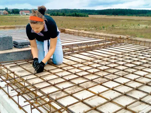 Kobieta w pomarańczowej opasce i jeansach klęczy, wiążąc zbrojenie. To element budowy domu samemu, gdzie każdy detal ma znaczenie.
